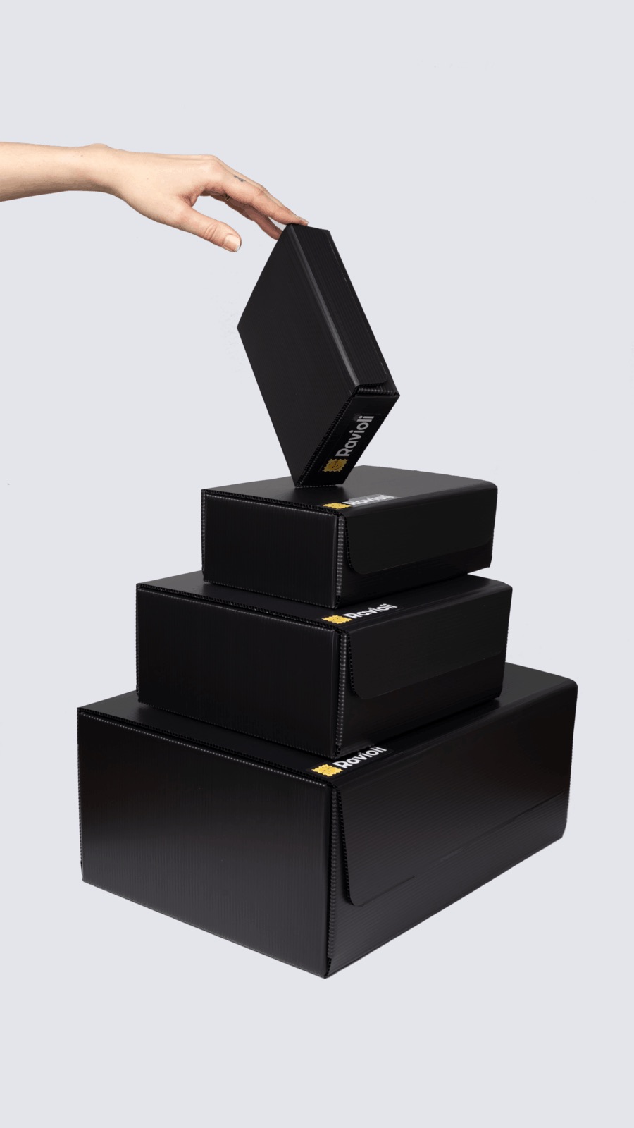 Vier anthrazitfarbene Ravioli Pakete sind vor hellgrauem Hintergrund aufeinander gestapelt. Eine Hand greift sich das oberste Paket.